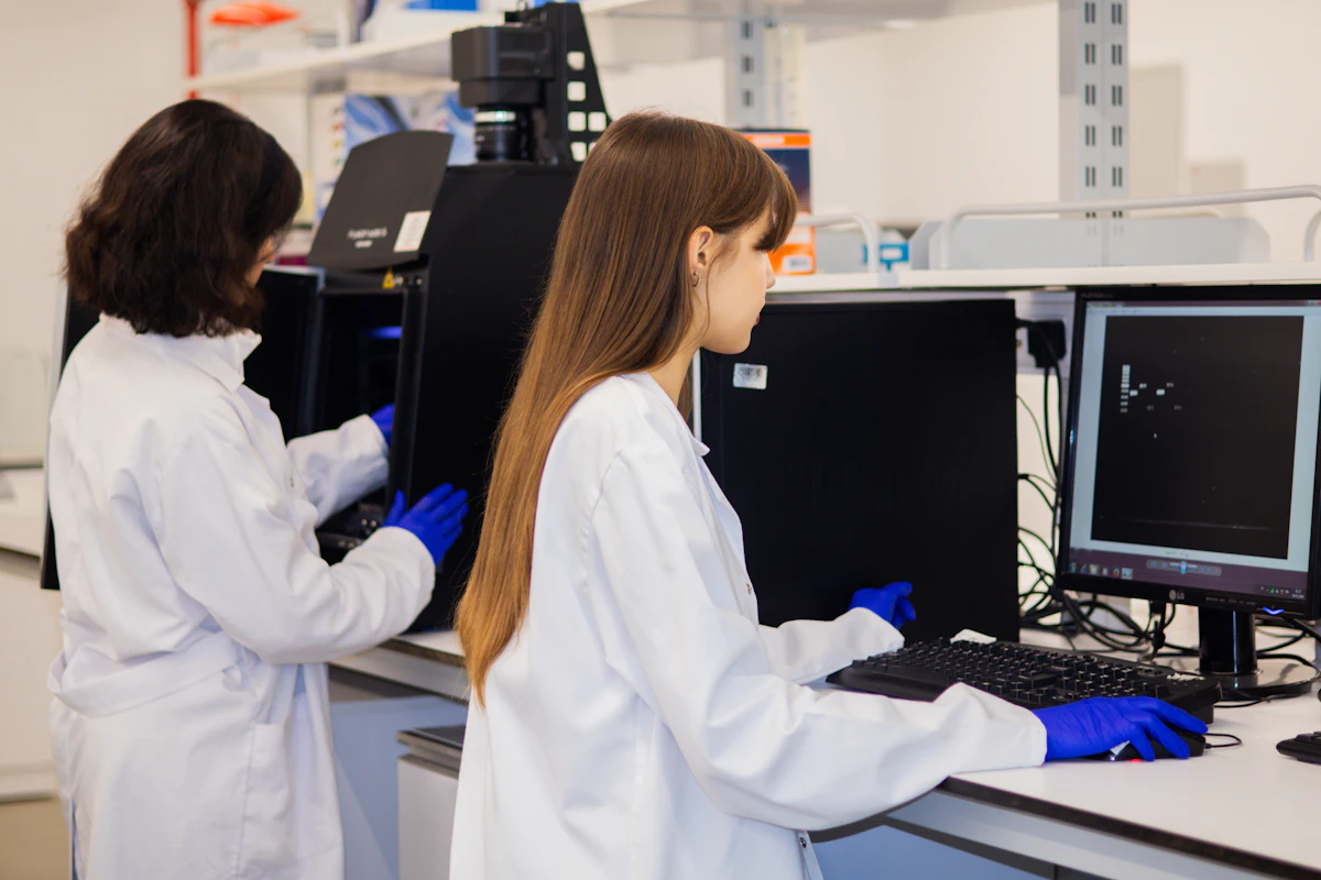 Dos investigadoras con bata y guantes trabajan junto a un instrumento de laboratorio y un ordenador